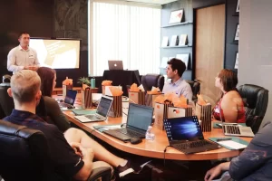 people sitting around a oval desk on their laptops, watching a presentation
