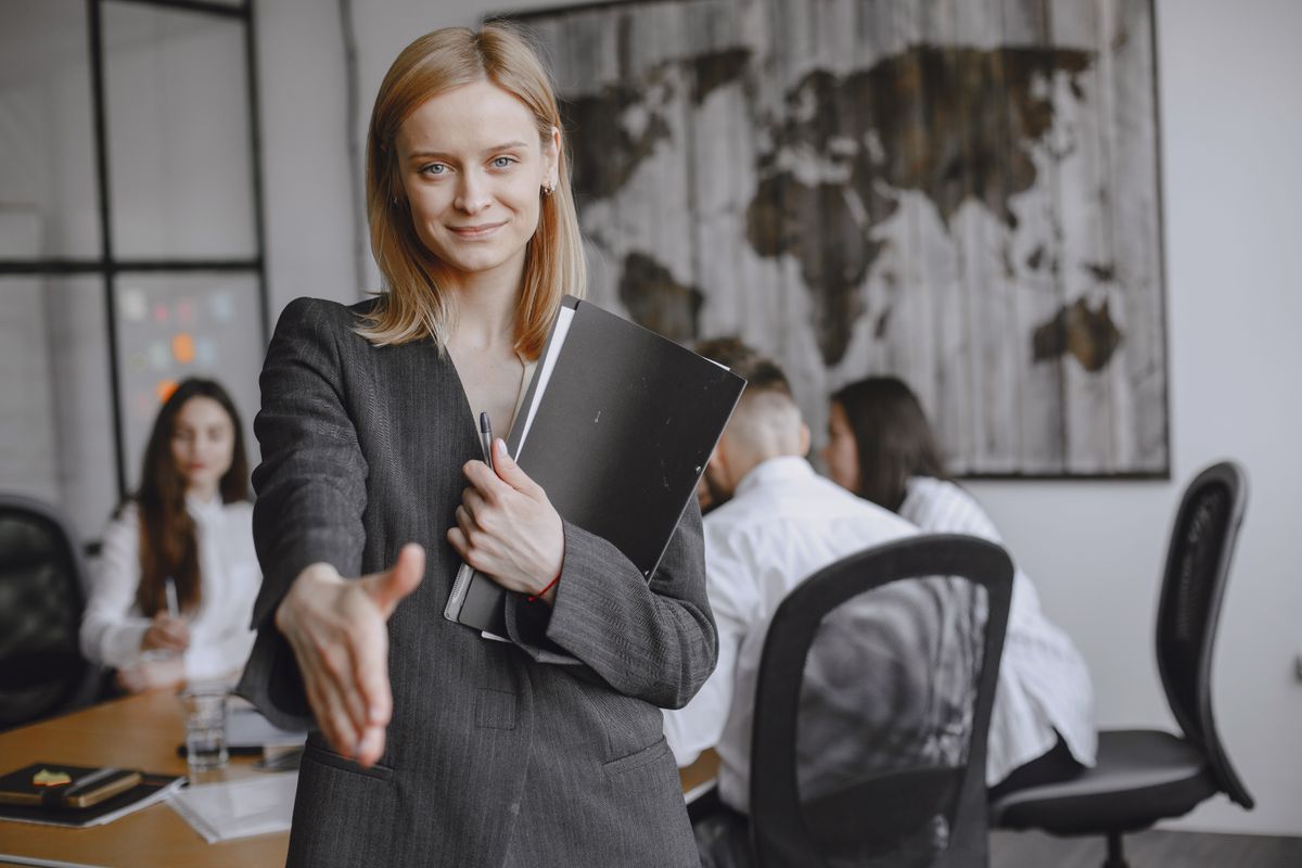 A lady holding her hand out to shake another person's hand in an office room filled with people